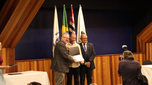 Fotografia que mostra três homens em pé em um palco, um ao lado do outro, sorrindo e olhando para uma câmera fotográfica. Um deles segura um prêmio. O ambiente é fechado e bem iluminado, há bandeiras, a parede é azul escuro e há uma mesa de madeira em cima do palco. Fim da descrição.